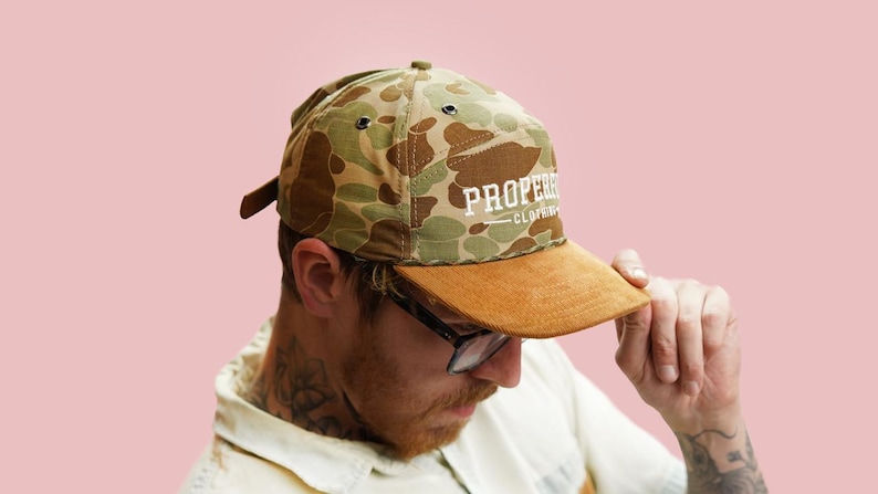 Puede incluir: Gorra de b&eacute;isbol con estampado de camuflaje y visera de ante marr&oacute;n. La gorra tiene el texto "PROPER CLOTHING" en letras blancas. El fondo es rosa claro liso.
