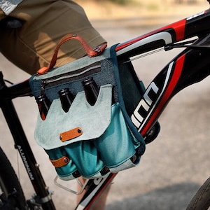 Puede incluir: Una bolsa de bicicleta de lona azul y turquesa con asa y correas de cuero. La bolsa está unida al cuadro de una bicicleta negra y contiene cuatro botellas de vidrio marrón.