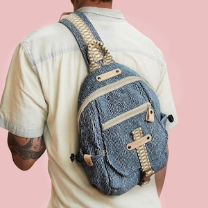 Puede incluir: Mochila bandolera de mezclilla azul con correa y detalles beige. La bolsa tiene un compartimento principal con cremallera y un bolsillo frontal más pequeño. La correa es ajustable y acolchada. La bolsa se lleva sobre el hombro.