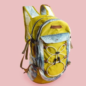 Puede incluir: Una mochila amarilla y azul con cierre de cord&oacute;n blanco y un parche de cuero marr&oacute;n. La mochila tiene varios bolsillos y correas.