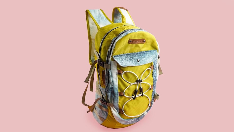 Puede incluir: Una mochila amarilla y azul con cierre de cord&oacute;n blanco y un parche de cuero marr&oacute;n. La mochila tiene varios bolsillos y correas.