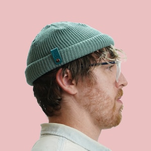 Könnte beinhalten: Ein Mann trägt eine tealfarbene Mütze mit einem Lederpatch und einer klaren Brille. Die Mütze ist gerippt und hat einen umgeschlagenen Rand.