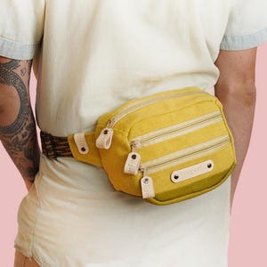 Op de afbeelding: Mosterdgele heuptas met drie vakken met ritssluiting en een bruin en tan patroon riem. De tas heeft leren accenten en wordt om de taille gedragen. De achtergrond is effen lichtroze.