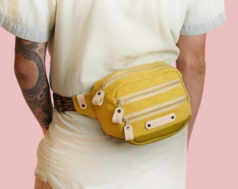 May include: Mustard yellow fanny pack with three zippered compartments and a brown and tan patterned belt. The bag has leather accents and is worn around the waist. The background is a solid light pink.