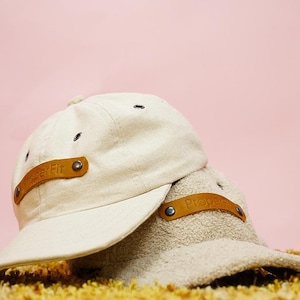 May include: Two beige baseball caps with brown leather straps. The straps have the word "Proper" embossed on them.