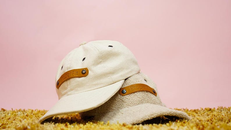 May include: Two beige baseball caps with brown leather straps. The straps have the word "Proper" embossed on them.