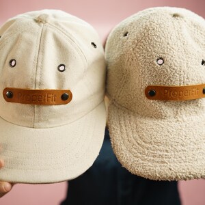 May include: Two beige baseball caps with brown leather patches that say "Proper Fit" on them. One cap is made of a smooth fabric and the other is made of a fuzzy fabric.