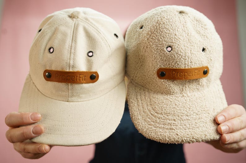 May include: Two beige baseball caps with brown leather patches that say "Proper Fit" on them. One cap is made of a smooth fabric and the other is made of a fuzzy fabric.