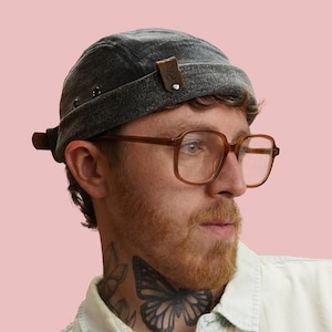 May include: A gray denim cap with brown leather accents and rivets. The cap is worn by a person with brown-framed glasses and a beard. The background is a solid pink color.