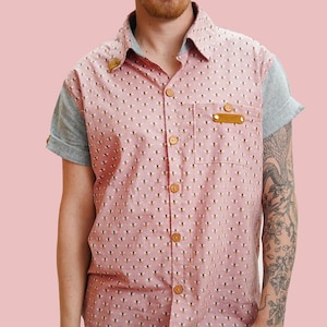 Könnte beinhalten: Ein rosa Hemd mit Knöpfen, einem weißen Muster und einem kleinen Lederflicken auf der Brust. Das Hemd hat kurze Ärmel und ist aus einem leichten Stoff gefertigt.