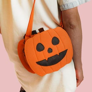 Puede incluir: Un bolso bandolera con forma de calabaza naranja con un diseño de cara negra. El bolso tiene una larga correa naranja y un pequeño detalle de tallo. El bolso está hecho de una tela texturizada y se lleva al hombro.