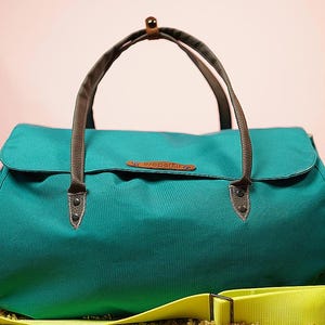 Puede incluir: Bolso de lona turquesa con asas marrones y una correa para el hombro de color amarillo brillante. El bolso tiene un cierre de solapa y una pequeña etiqueta de cuero. El bolso está sobre una superficie texturizada con un fondo rosa.