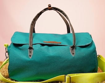 Puede incluir: Bolso de lona turquesa con asas marrones y una correa para el hombro de color amarillo brillante. El bolso tiene un cierre de solapa y una pequeña etiqueta de cuero. El bolso está sobre una superficie texturizada con un fondo rosa.