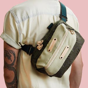 Peut inclure: Sac bandoulière vert et beige avec une sangle noire. Le sac, porté sur l'épaule, est de forme rectangulaire avec un compartiment zippé et des détails en cuir. Le texte "Proper Fit" est visible.
