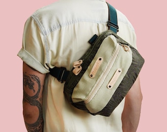 May include: A green and beige crossbody bag with a black strap is worn over the shoulder. The bag has a rectangular shape with a zippered compartment and leather accents. The text "Proper Fit" is visible on the bag.