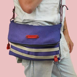 Puede incluir: Un bolso bandolera de lona morado con una correa de cuero roja y un detalle de rayas blancas y grises. El bolso tiene cierre de solapa y una pequeña etiqueta de cuero con la palabra "WAKAO" en ella.