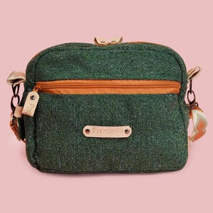 May include: Green crossbody bag with an orange zipper and strap accents. The bag has a small leather tag and a label that reads "ProdeFit". The bag is set against a pink background.