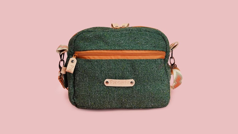 May include: Green crossbody bag with an orange zipper and strap accents. The bag has a small leather tag and a label that reads "ProdeFit". The bag is set against a pink background.