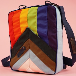 Op de afbeelding: Een regenboog gestreepte schoudertas met een chevron-patroon aan de voorkant. De tas heeft een donkerblauwe band met een bruin leren label waarop "Doodad" staat.