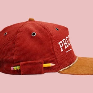Pu&ograve; includere: Un berretto da baseball in velluto a coste rosso con visiera marrone e un logo ricamato bianco che dice "PRO" con una linea sottostante. Una matita gialla &egrave; infilata in una tasca sul lato del berretto.