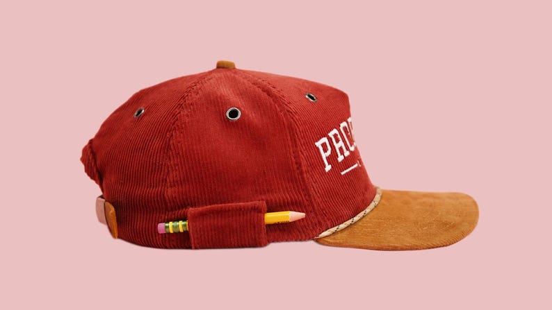 Pu&ograve; includere: Un berretto da baseball in velluto a coste rosso con visiera marrone e un logo ricamato bianco che dice "PRO" con una linea sottostante. Una matita gialla &egrave; infilata in una tasca sul lato del berretto.