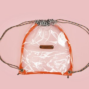 May include: Clear drawstring backpack with orange trim and brown and black rope straps. The bag has a small brown leather tag and a black and white checkered fabric detail at the top.