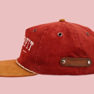 Pu&ograve; includere: Un cappello da baseball in velluto a coste rosso con visiera in pelle scamosciata marrone. Il cappello ha un logo ricamato bianco che dice "Pre-Fit" e una toppa in pelle marrone che dice "Preperfkt".