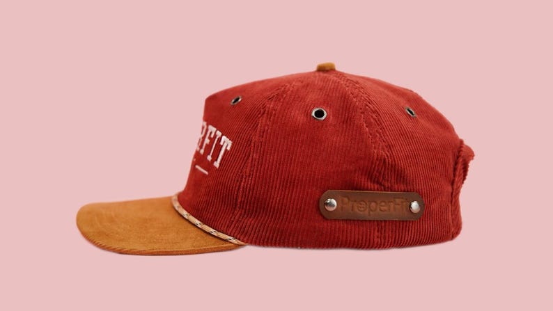 Pu&ograve; includere: Un cappello da baseball in velluto a coste rosso con visiera in pelle scamosciata marrone. Il cappello ha un logo ricamato bianco che dice "Pre-Fit" e una toppa in pelle marrone che dice "Preperfkt".