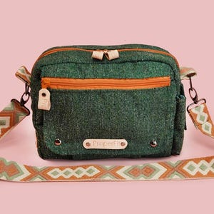 May include: Green crossbody bag with an orange zipper and strap accents. The bag has a patterned strap with geometric designs in shades of orange, white, and green. The bag has the text "ProperFit" on the front.