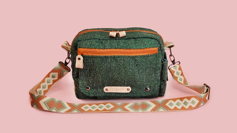 May include: Green crossbody bag with an orange zipper and strap accents. The bag has a patterned strap with geometric designs in shades of orange, white, and green. The bag has the text "ProperFit" on the front.