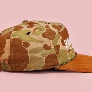 Puede incluir: Gorra de b&eacute;isbol con estampado de camuflaje con visera de pana marr&oacute;n y correa de cuero marr&oacute;n. La gorra presenta la palabra "PRO" en letras blancas. El fondo es de color rosa liso.