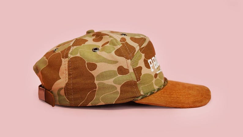 Puede incluir: Gorra de b&eacute;isbol con estampado de camuflaje con visera de pana marr&oacute;n y correa de cuero marr&oacute;n. La gorra presenta la palabra "PRO" en letras blancas. El fondo es de color rosa liso.