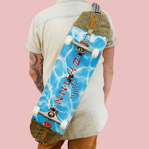 May include: A person wearing a light-coloured shirt is carrying a blue and white skateboard with a water-like pattern on the deck. The skateboard is secured to a brown and black strap.