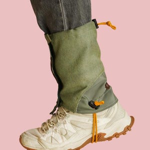 Könnte beinhalten: Eine olivgrüne Gamasche über einem weißen Sportschuh. Die Gamasche verfügt über einen vertikalen Reißverschluss, einen verstellbaren orangefarbenen Kordelzug am oberen Rand und eine passende orangefarbene Schnur unter der Sohle.