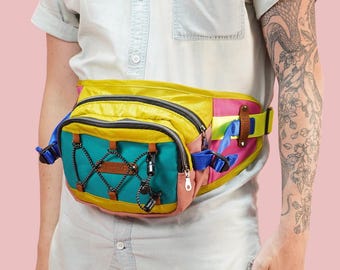 Puede incluir: Una riñonera multicolor con un diseño amarillo, rosa, azul y turquesa. La riñonera tiene varios compartimentos y un cierre de hebilla.