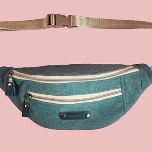 K&ouml;nnte beinhalten: Eine t&uuml;rkisfarbene Bauchtasche mit zwei Rei&szlig;verschlussf&auml;chern und einem verstellbaren beigefarbenen Gurt. Die Tasche hat ein kleines wei&szlig;es Etikett mit dem aufgedruckten Wort "Stonemill".