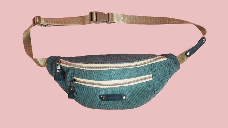 K&ouml;nnte beinhalten: Eine t&uuml;rkisfarbene Bauchtasche mit zwei Rei&szlig;verschlussf&auml;chern und einem verstellbaren beigefarbenen Gurt. Die Tasche hat ein kleines wei&szlig;es Etikett mit dem aufgedruckten Wort "Stonemill".