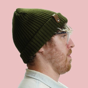 Puede incluir: Un hombre con una boina verde con un parche de cuero marrón y gafas de montura transparente. Está mirando hacia la derecha.