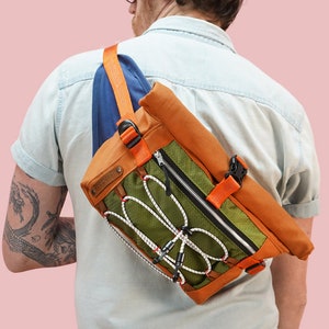 May include: A person wearing a light blue shirt is wearing a green, orange, and blue crossbody bag with a zipper and a buckle closure.