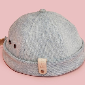 May include: A light gray denim cap with a short brim and a small, rounded top. The cap features a brown leather strap on the side and a small tag with text. The background is a solid pink.