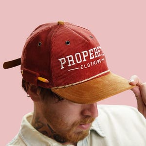 Könnte beinhalten: Eine rote Baseballkappe aus Cord mit braunem Schirm und einem weißen gestickten Logo, auf dem "PROPER" mit "CLOTHING" darunter steht. Die Kappe hat einen Stifthalter an der Seite.