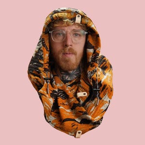May include: A hooded scarf with a vibrant orange and black floral pattern. The scarf has a toggle closure and is worn around the neck. The background is a solid light pink.