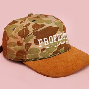 May include: A camouflage pattern cap with a brown corduroy brim. The cap features the text "PROPERFIT CLOTHING" in white block letters. The background is a solid light pink.