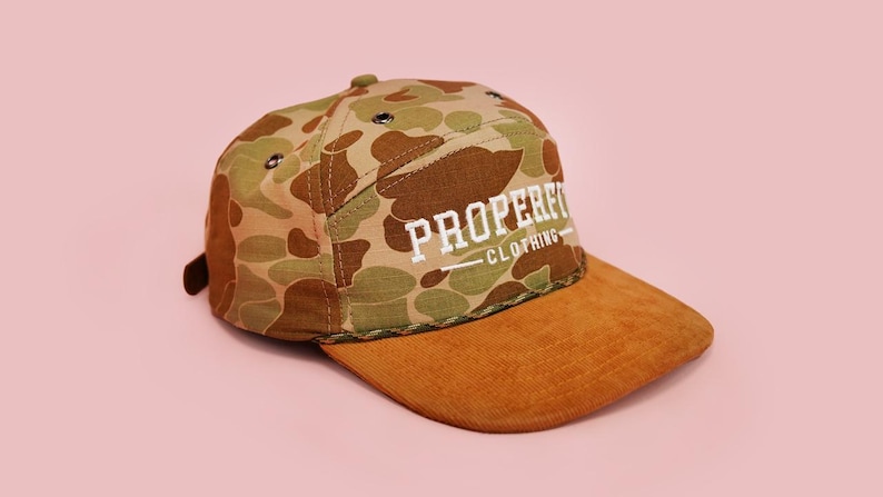 Puede incluir: Gorra con estampado de camuflaje y visera de pana marr&oacute;n. La gorra presenta el texto "PROPERFIT CLOTHING" en letras blancas. El fondo es de color rosa claro liso.