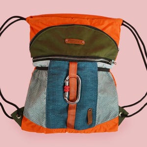 May include: A colorful drawstring backpack with orange, green, and blue panels. The bag features a large silver carabiner clip and black drawstrings. The design includes a mesh pocket and a small leather tag.
