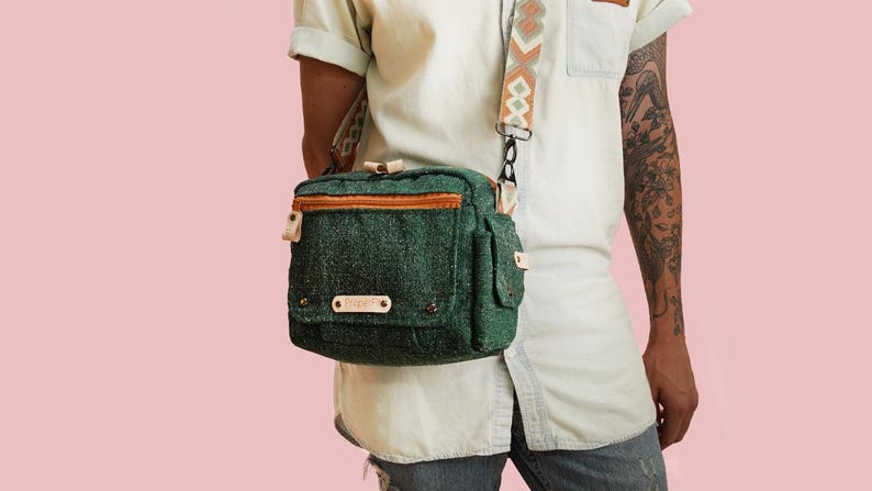 May include: A green crossbody bag with a textured fabric, featuring a brown zipper and strap accents. The bag has a patterned strap and a small label with the word "Prophet".