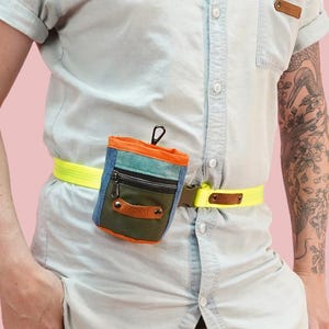 Puede incluir: Una riñonera multicolor con un cinturón amarillo neón y una pequeña bolsa. La bolsa tiene un frente azul vaquero, un cuerpo verde oliva y una parte superior naranja. El cinturón tiene un acento de cuero marrón.