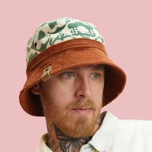 Puede incluir: Un sombrero de pescador de pana con un estampado floral verde y blanco en la corona y una banda marrón. El sombrero tiene un cierre metálico y la palabra "PROPHECY" impresa en la corona. El fondo es rosa liso.