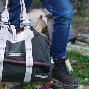 May include: A green canvas and leather tote bag with a brown axe secured on the outside. The bag has white leather straps and accents. The person is wearing blue jeans and brown boots.