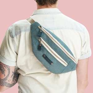 以下が含まれることがあります： 白のストライプと2つのジッパーが付いたターコイズブルーのウエストバッグ。ウエストバッグは、薄いブルーのシャツを着た人の体にかけられています。